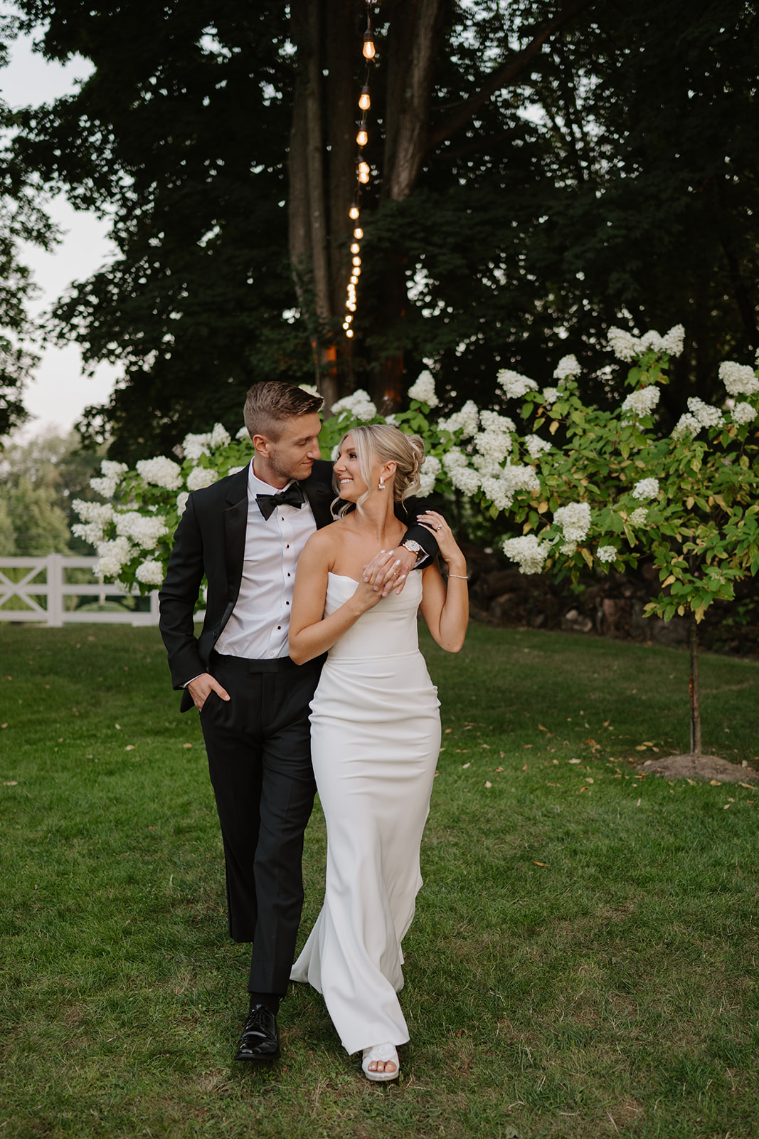 Newlyweds walk hand-in-hand on the grass, smiling at each other by blooming white hydrangeas.